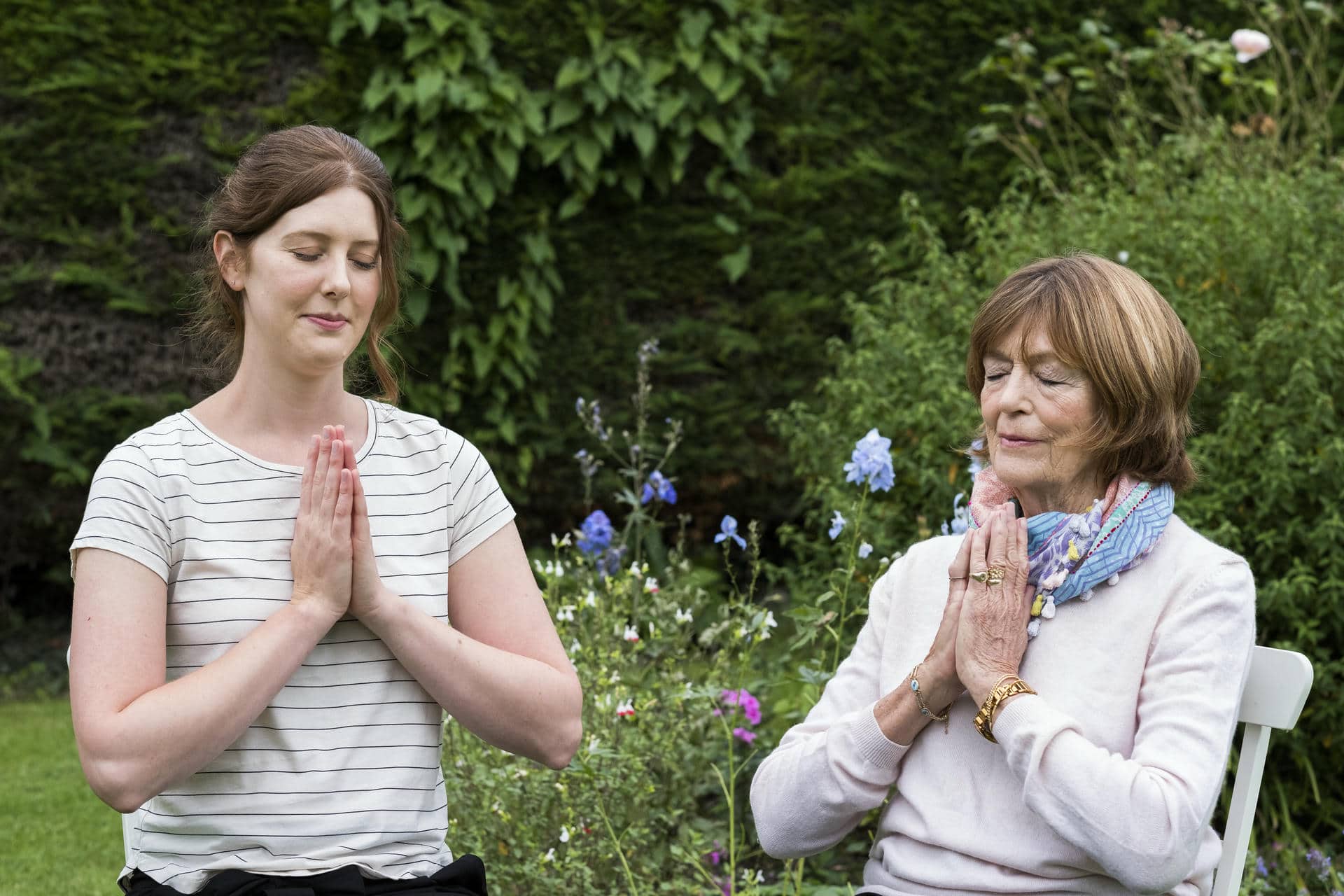 Woman and female therapist seated in a garden, hands together and eyes closed.